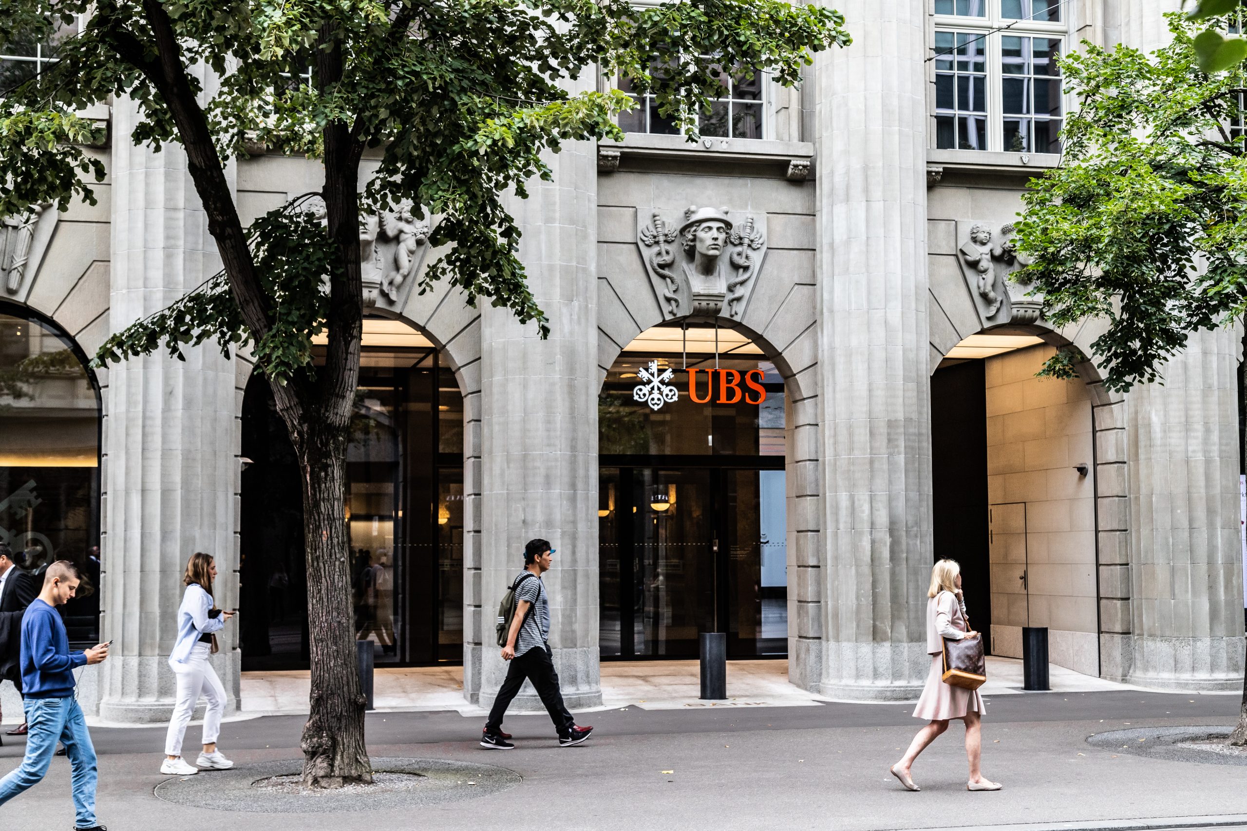 A street-level view of a UBS bank branch, with pedestrians walking past.