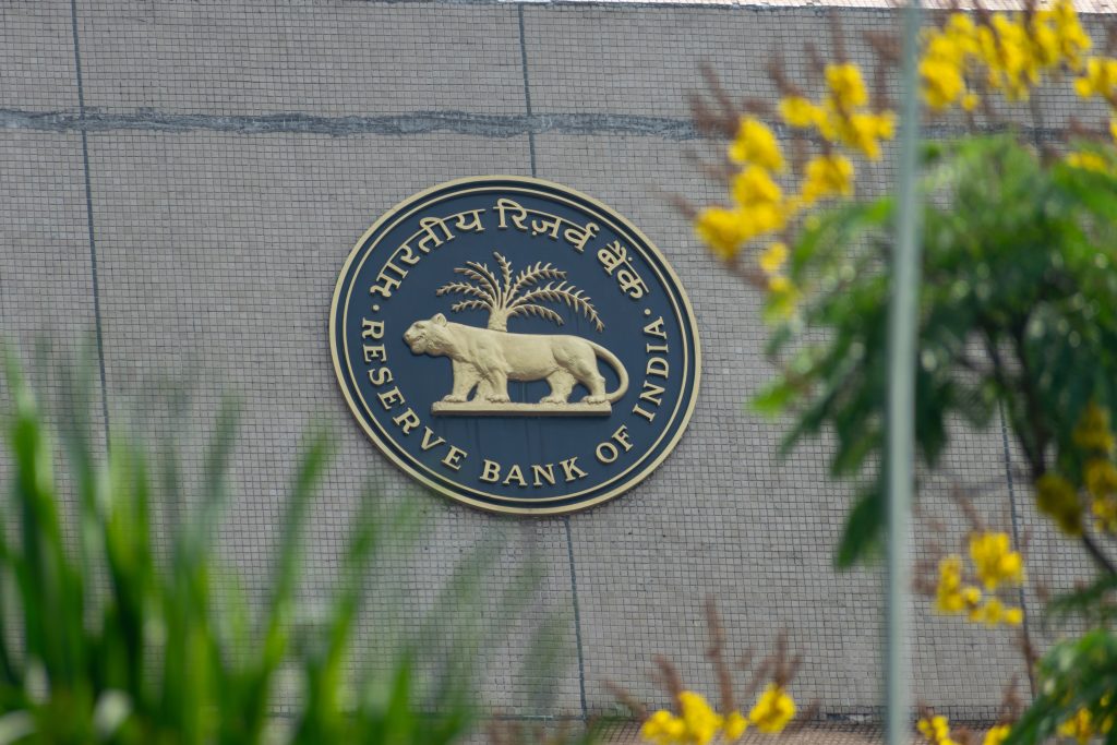 The emblem of the Reserve Bank of India on a building, partially obscured by green foliage and yellow flowers, representing the central bank's role in regulating India's financial system amidst economic changes.