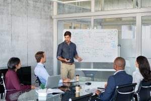 A man leads a presentation in a modern conference room, gesturing towards a whiteboard filled with notes while a diverse team of colleagues listens. The scene represents a crucial stage of business development, such as strategic planning or a pitch, illustrating the growth journey from freelancer to a larger company as discussed in the article.