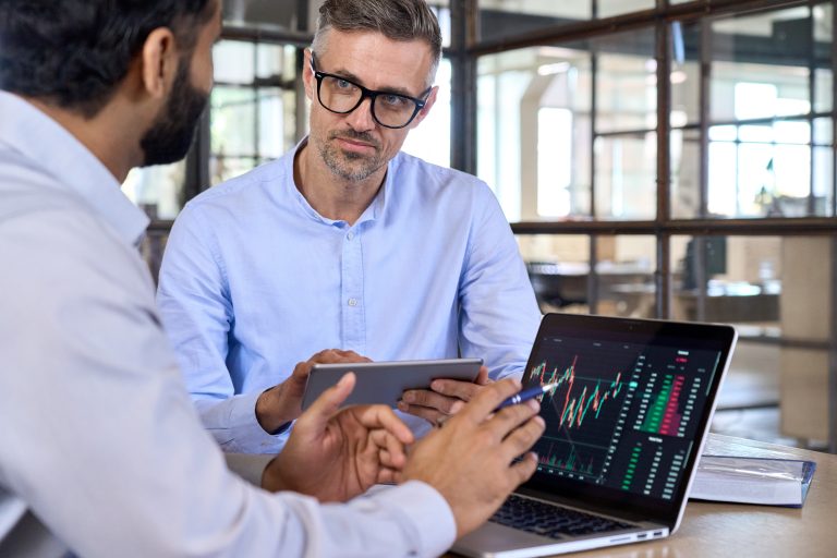 Two professionals in an office analyze a financial candlestick chart on a laptop. The scene represents the collaborative and tech-driven approach of modern financial advice, as discussed in the 2025 article about new models challenging traditional banks.