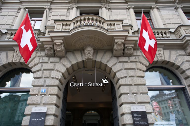 The entrance to a historic Credit Suisse bank building, flanked by two Swiss flags. The image of a major Swiss financial institution represents the country's robust financial sector, which is the focus of a 2025 article about its booming bond market.