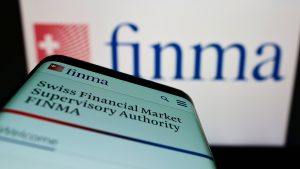 A smartphone displays the official website of the Swiss Financial Market Supervisory Authority (FINMA). In the background, the FINMA logo is prominently shown on another screen. The image represents the Swiss financial regulator, which is the subject of a 2025 article about its new supervisory approach.
