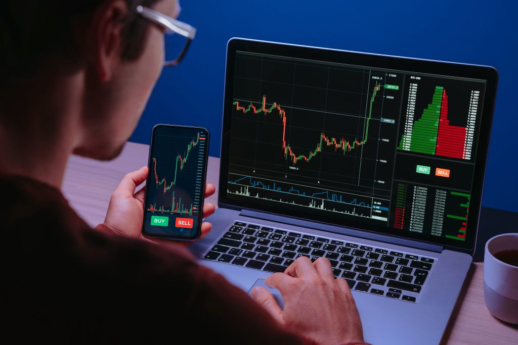 A trader analyzes cryptocurrency charts on both a laptop and a smartphone, with "BUY" and "SELL" buttons visible on the phone. The image represents the act of crypto trading, which companies like Cryptonow are bringing into the regulated "mainstream finance" of the EU, as discussed in the October 2025 article about their new MiCAR license.