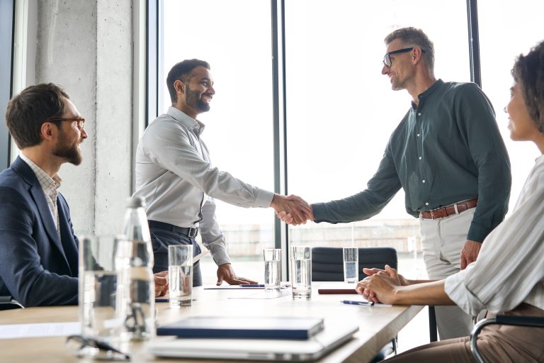 Two businessmen shake hands across a conference table, symbolizing a new partnership. The image illustrates the agreement mentioned in the October 2025 article about Bank Frick adopting the GenTwo platform to expand its digital asset capabilities.
