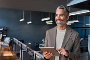 A smiling, mature executive with gray hair holding a tablet in a modern office, representing the Warburg Pincus veteran tapped by Partners Group to co-lead its technology investments.