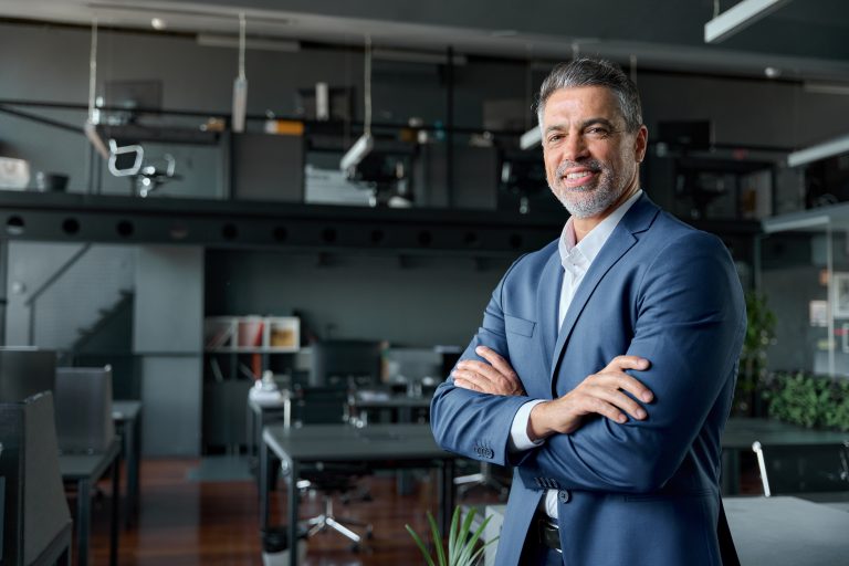 A confident, smiling senior executive in a suit stands with arms crossed in a modern office. This image represents former SNB President Thomas Jordan, who has joined Avenir Suisse as a Distinguished Fellow.