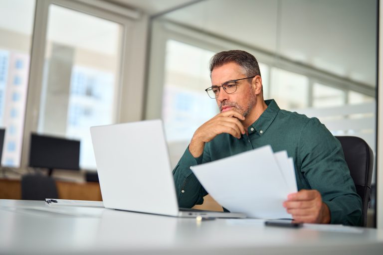 A focused investor analyzing documents and data on a laptop in a modern office setting.